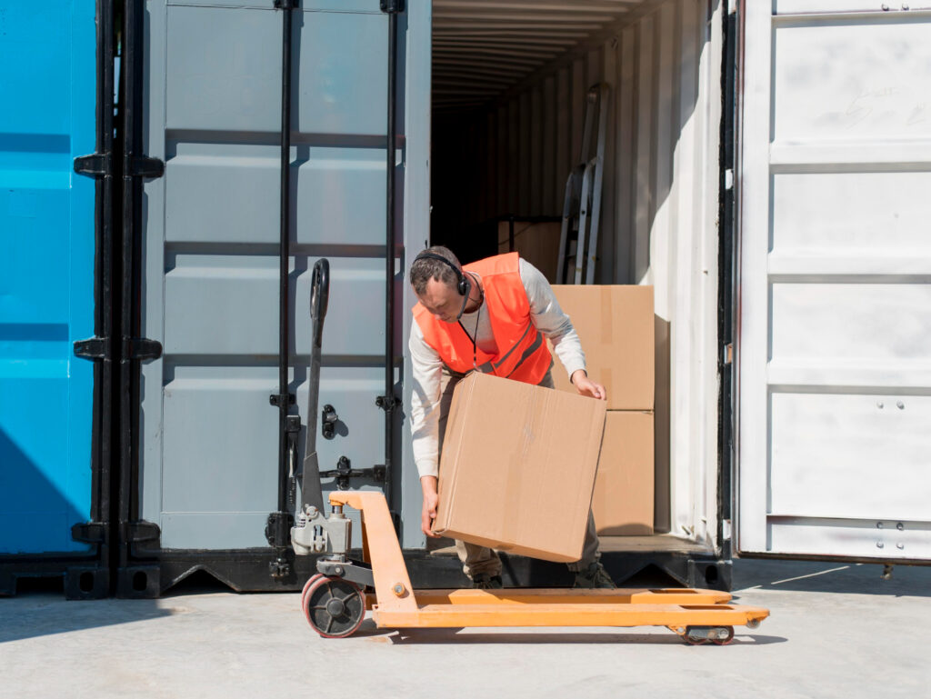 Worker in orange vest unloading cardboard boxes from a shipping container using a pallet jack