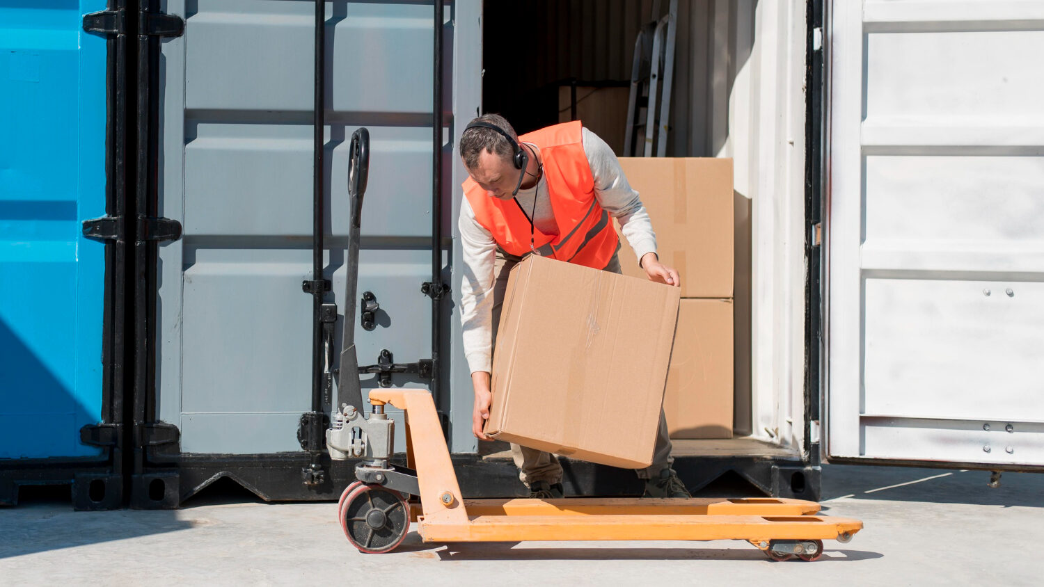 Worker in orange vest unloading cardboard boxes from a shipping container using a pallet jack